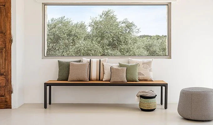 Wooden bench with cushions against a wall with a large window showing greenery outside.