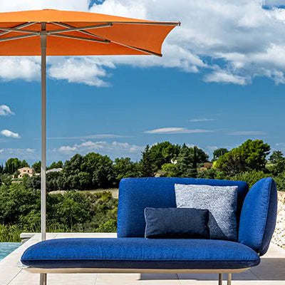 Blue outdoor lounge chair with orange umbrella by a poolside with greenery and blue sky.