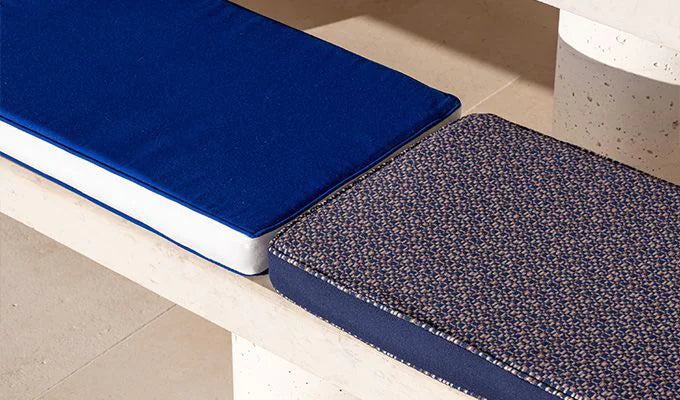 Blue cushion on a white bench next to a textured blue rug on a beige floor.