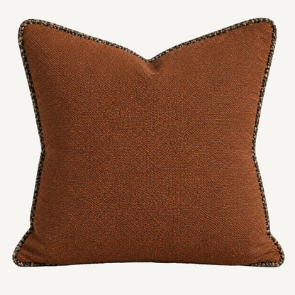A square decorative throw pillow in a textured rust-brown fabric with a dark gray and beige braided trim, shown against a neutral beige background.